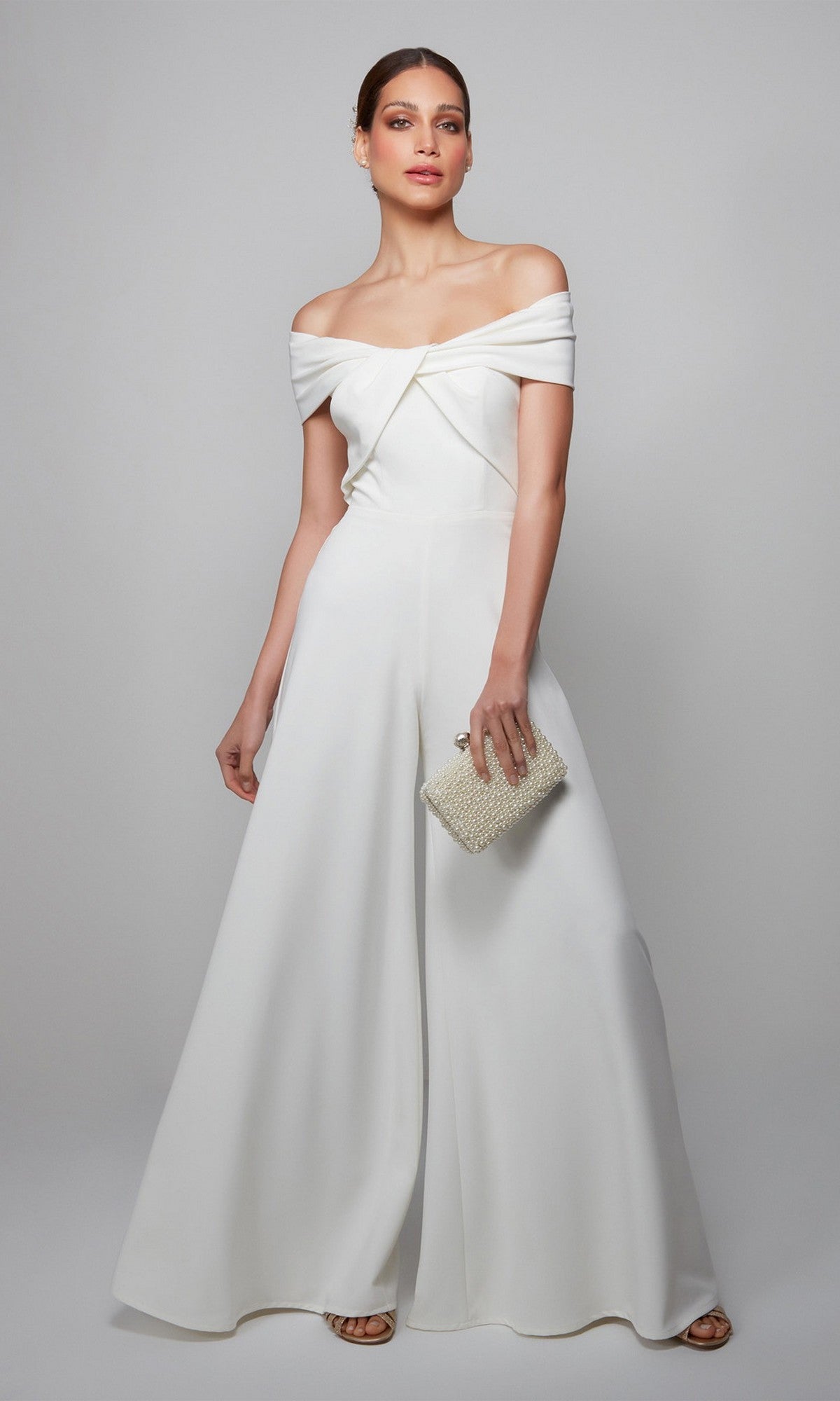 A woman stands against a plain background wearing the Alyce 70013 long formal jumpsuit, paired with a beaded clutch and sandals.